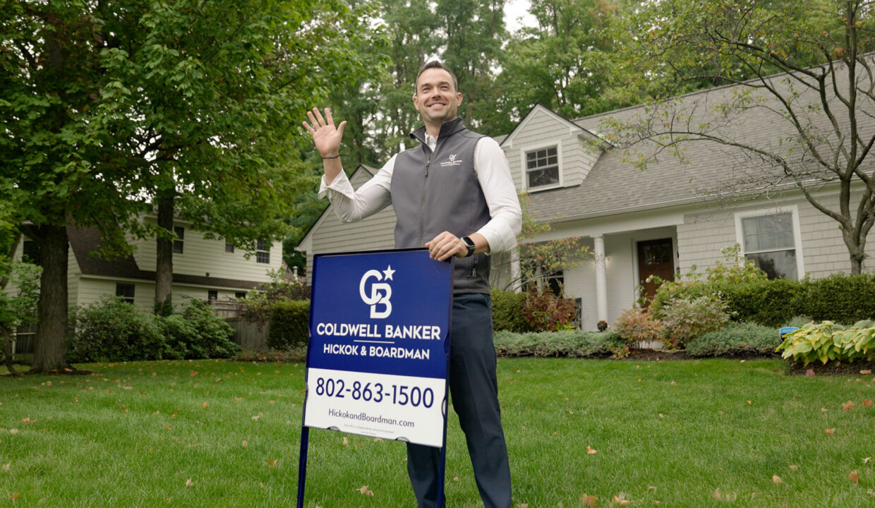 Realtor® John Black standing in front of a residential home, holding a Coldwell Banker Hickok & Boardman yard sign and waving to greet clients