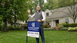 Realtor® John Black standing in front of a residential home, holding a Coldwell Banker Hickok & Boardman yard sign and waving to greet clients