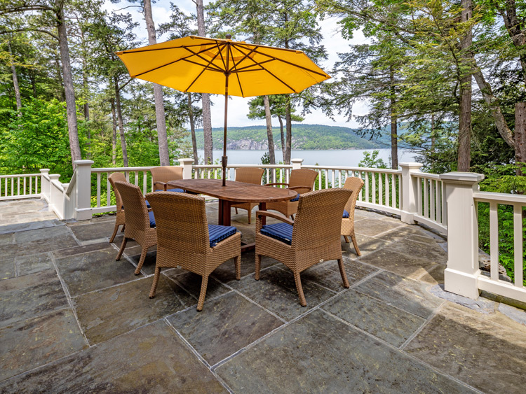 Stone terrace with outdoor dining table and umbrella overlooking Lake Champlain, surrounded by mature trees and scenic waterfront views.