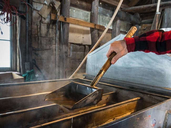A person in a red plaid shirt skims steaming maple syrup inside a sugarhouse, with stainless steel evaporator pans and rustic wooden beams visible, reflecting Vermont’s spring sugaring season in St. Albans.