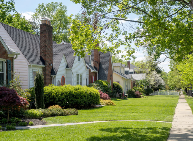 Tree-lined neighborhood street with classic single-family homes, brick chimneys, manicured lawns, and a wide sidewalk on a sunny day.