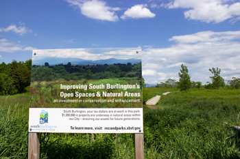 Sign in a South Burlington natural area highlighting investment in open space and conservation, with a grassy field, walking path, and distant Green Mountain views under a blue sky with scattered clouds.